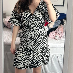 Zebramönstrad klänning - Snygg zebramönstrad klänning med omlott-design och korta ärmar. Perfekt för en trendig look med sin v-ringade stil och volangdetaljer vid fållen. En riktig statement-piece för garderoben!