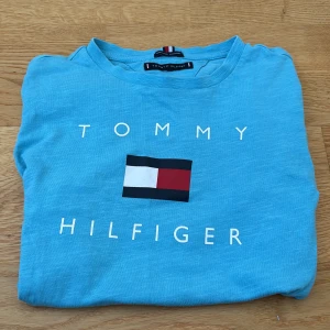 Blå t-shirt från Tommy Hilfiger - Snygg blå t-shirt från Tommy Hilfiger i ekologisk bomull. Den har korta ärmar och ett stort tryck med logotypen på framsidan. Perfekt för en avslappnad stil.