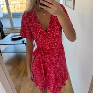 Röd kortklänning med vita prickar - Charmig röd kortklänning med vita prickar och omlott-design. Klänningen har korta ärmar och en knytning i midjan som ger en smickrande passform. Perfekt för sommarens varma dagar. Använd endast 1 gång!
