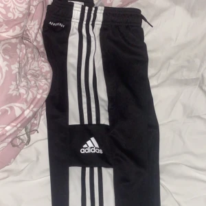 Svarta träningsbyxor från Adidas - Säljer ett par svarta träningsbyxor från Adidas med vita ränder längs sidorna och logotyp på benet. Byxorna har en elastisk midja med snörning för justerbar passform. Perfekta för sport och träning!