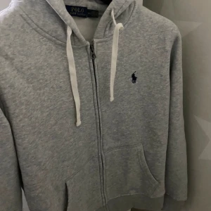Grå hoodie från Polo Ralph Lauren - Säljer en stilren grå hoodie från Polo Ralph Lauren med dragkedja och justerbar huva. Den har en broderad logga på bröstet och praktiska fickor framtill. Perfekt för en avslappnad look.