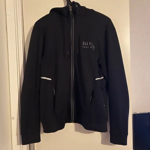 Svart hoodie från Hugo Boss - Säljer en stilren svart hoodie från Hugo Boss med dragkedja och vita detaljer. Perfekt för en avslappnad look. Den har långa ärmar och en praktisk huva.