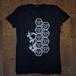 Svart Philipp Plein T-Shirt - Snygg Philipp Plein T-shirt i mycket bra skick! Svart bas med stilren design: vita hexagonloggor med “qp”-mönster och ett halvt dödskallemotiv som ger tröjan en unik edge. Perfekt för dig som vill sticka ut med lyxig streetwear. Materialet är mjukt och stretchigt – både bekvämt och hållbart. Passar lika bra till vardags som till fest. Storlek: S
