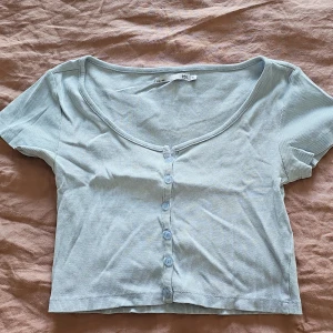 Ljusblå ribbad topp från 157 - Säljer en ljusblå ribbad topp från 157 med korta ärmar och knappar framtill. Perfekt för en avslappnad stil. Toppen har en rundad halsringning och är i en mjuk och bekväm bomullsmix.
