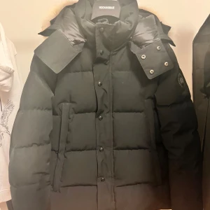 Canada Goose Wyndham Black label - Säljer en svart dunjacka från Canada Goose med avtagbar huva och pälsdetalj. Jackan har långa ärmar och stängs med dragkedja och knappar. Perfekt för kalla vinterdagar.                                Storlek M, kontakta i Pm för mer info