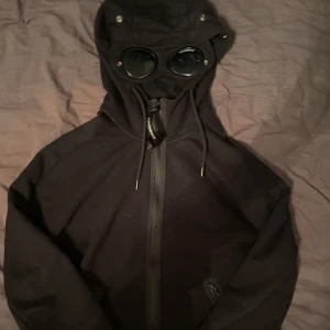 Svart hoodie från C.P. Company - Säljer en svart hoodie från C.P. Company med inbyggda glasögon i huvan. Hoodien har dragkedja framtill och är långärmad. Perfekt för en unik och stilren look. Köptes för 3400 kr i Halmstad.