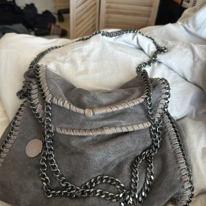 Stella macartney liknande väska - Snygg grå axelväska med kedjedetaljer och dekorativa sömmar. Väskan har en kedjerem och en rymlig insida med röd foder. Perfekt för att ge din outfit en cool touch.