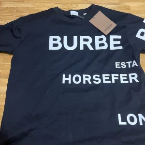 Svart Burberry T-shirt - Snygg svart t-shirt från Burberry med vit text på både fram- och baksidan. Perfekt för en avslappnad stil med en touch av lyx. T-shirten har en klassisk passform och korta ärmar. Den är rätt så oversized i storlek passar L-XL