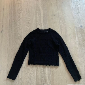 Svart stickad tröja från VERO MODA - Säljer en svart ribbad tröja från VERO MODA. Tröjan har långa ärmar och en fin struktur med vågiga kanter vid ärmslut och nederkant. Perfekt för en stilren look.