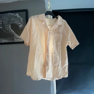 Beige kortärmad skjorta - Säljer en stilren beige kortärmad skjorta med knappar framtill. Skjortan har en klassisk krage och en bröstficka. Perfekt för varma dagar. Använd fåtal gånger, hör av dig vid fler bilder el funderingar!