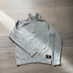 Beige stickad tröja från Calvin Klein - Säljer en stilren beige stickad tröja från Calvin Klein med halv dragkedja och hög krage. Tröjan har långa ärmar och ribbade muddar. Perfekt för våren. 🧥