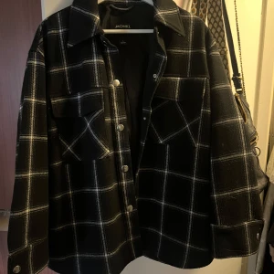 Svart och vit rutig jacka från Monki - Snygg svart och vit rutig jacka från Monki med knappar framtill. Jackan har en klassisk krage och två bröstfickor. Perfekt för en stilren look.