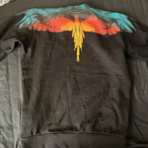 Svart tröja med färgglatt vingmönster från Marcelo Burlon - Säljer en svart tröja från Marcelo Burlon med ett iögonfallande vingmönster i blått, rött och gult över axlarna. Perfekt för dig som vill sticka ut med en unik design. Tröjan har långa ärmar och en rund halsringning. Ny pris 4 till 5 tusen 