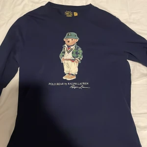 Mörkblå långärmad tröja från Ralph Lauren - Säljer en snygg mörkblå långärmad tröja från Ralph Lauren med en ikonisk Polo Bear-design på framsidan. Den är i topp skick och säljs för att den inte används. Den är i storlek xl för barn men passar s för vuxna 