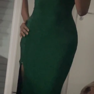 Grön elegant klänning - Säljer en helt ny och oanvänd lång klänning i en djup smaragdgrön färg med glittrigt tyg. Klänningen är kroppsnära med ett stretchigt material som formar sig snyggt efter kroppen och har en stilren slits framtill som ger en elegant men ändå sexig look.  Ryggen är öppen med korsade band som knyts, vilket ger en extra snygg detalj och gör att du kan justera passformen själv. Tunna axelband och figurnära siluett – perfekt för bal, gala, nyår eller andra festliga tillfällen där du vill sticka ut.  