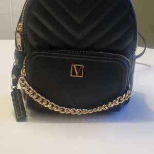 Svart ryggsäck från Victoria's Secret - Snygg svart ryggsäck från Victoria's Secret med quiltad design och gulddetaljer. Väskan har en kedja framtill och dragkedja för säker förvaring. Perfekt för att bära dina dagliga nödvändigheter med stil. Finns ej att köpa i Sverige. Är köpt i Polen på flygplatsen. Äkta 