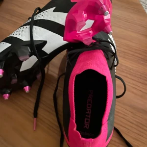 Adidas Predator fotbollsskor - Snygga Adidas Predator fotbollsskor i svart och rosa med vita detaljer. Skorna har snörning och en modern design med greppvänliga dobbar för optimalt fäste på planen. Perfekta för dig som vill dominera spelet med stil.