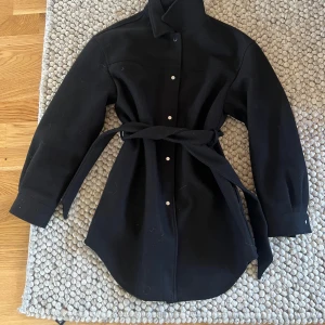 Svart kappa med bälte - Snygg svart kappa med knappar och bälte i midjan. Perfekt för höst och vår. Den har långa ärmar och en klassisk krage. Ifrån Vero Moda i storlek M