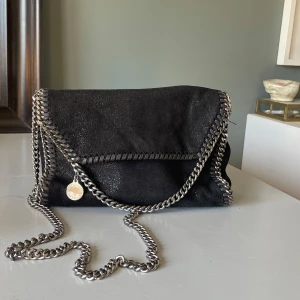 Svart axelväska från Stella McCartney - Snygg svart axelväska från Stella McCartney med kedjedetaljer och magnetlås. Väskan har en stilren design med en glittrig yta och en insida i rosa tyg med logotypmönster. Pris kan diskuteras!