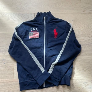 Mörkblå tröja från Polo Ralph Lauren - Säljer en mörkblå tröja från Polo Ralph Lauren med dragkedja och USA-flagga på bröstet. Jackan har långa ärmar med vita ränder längs sidorna och en röd logotyp på framsidan. Perfekt för en vårig look!