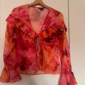 Blus  - Det här är en tunn, blommig blus från Boohoo i transparent tyg. Den har ett färgstarkt mönster i rosa, rött och orange toner. Blusen har en djup V-ringning med volanger längs bröstet och knytning framtill. Ärmarna är långa och vida med volanger vid ärmsluten. En feminin och somrig topp med en romantisk känsla. Typ helt ny och bara använt 1 gång 