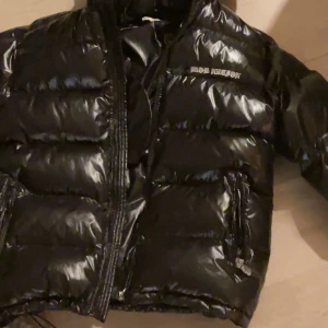 Svart pufferjacka från Mob Nation - Snygg svart pufferjacka från mob Nation med glansig finish. Jackan har en dragkedja framtill och är perfekt för kyligare dagar. Den har långa ärmar och praktiska fickor med dragkedja. Hör av er om ni har frågor. ✌️
