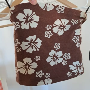 Tubetopp - Säljer en brun tubtopp från H&M Divided med vita blommor. Perfekt för sommardagar med sitt lätta och bekväma material☀️. Toppen är ärmlös och har ett tropiskt mönster som ger en avslappnad stil🌺. Den är i storlek XXS och använd fåtal gånger, pris diskuteras💗💗😘🎉
