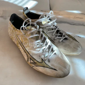 Vita fotbollsskor från Mizuno - Snygga vita fotbollsskor från Mizuno med gulddetaljer och snörning. Skorna har en rund tå och är designade för optimal prestanda på planen. Perfekta för dig som vill ha stil och funktion i ett. Inga skador på dem