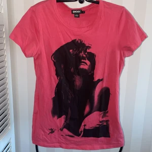 Rosa t-shirt från DKNY ny - Snygg rosa t-shirt från DKNY med ett coolt tryck av en kvinna på framsidan. Perfekt för en avslappnad stil med en touch av attityd. Kortärmad och tillverkad i mjukt material för extra komfort.