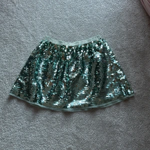 Grön paljettkjol från H&M - Snygg grön kjol från H&M täckt med glittrande paljetter. Perfekt för att lysa upp vilken outfit som helst. Kjolen har en elastisk midja för bekväm passform.