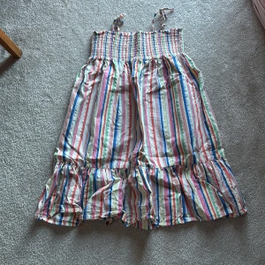 Randig strandklänning från H&M - Säljer en färgglad randig klänning från H&M med smockdetaljer upptill och volang nedtill. Klänningen har knutna axelband och är perfekt för sommarens varma dagar. Materialet är mestadels bomull med en liten del metalliserad fiber för extra glans.