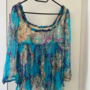 Blommig långärmad topp i blått och lila - Säljer en vacker blommig topp med långa ärmar i en mix av blått, lila och beige. Toppen har en luftig passform och ett romantiskt mönster med volangdetaljer vid halsringningen. Perfekt för en bohemisk look!