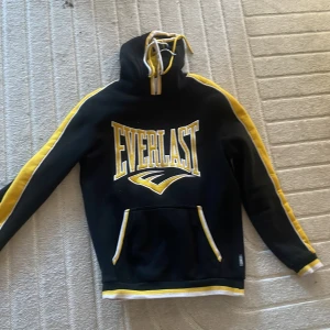 Svart och gul hoodie från Everlast - Säljer en svart hoodie från Everlast med gula detaljer och text. Den har en stor ficka framtill och en justerbar huva med snören. Perfekt för en sportig look!