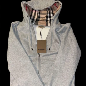 Grå hoodie från Burberry - Säljer en stilren grå hoodie från Burberry med klassiskt rutigt mönster i huvan. Den har dragkedja framtill och snörning vid huvan. Perfekt för en avslappnad och trendig look.