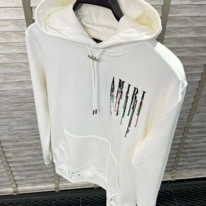 Vit hoodie från Amiri - Säljer en stilren vit hoodie från Amiri med färgglada detaljer som rinner ner från logotypen. Hoodien har en klassisk passform med känguruficka och justerbar dragsko i huvan. Perfekt för en avslappnad look.