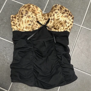 Leopardmönstrad korsettopp - Snygg korsettopp med leopardmönster upptill och svart rynkad design nedtill. Toppen är axelbandslös och har en  rosett framtill. 