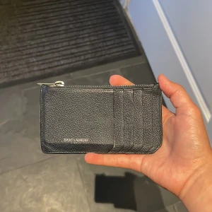 ÄKTA saint Laurent plånbok - Slimmad svart plånbok från Saint Laurent i äkta skinn med silvrig dragkedja och flera kortfack på framsidan. Perfekt för att hålla koll på kort och kontanter i en stilren och lyxig design. Diskret logga framtill. Värde över 6000kr!