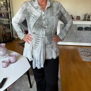 Grå skjorta från Elisa Cavaletti - Unik grå skjorta från Elisa Cavaletti med coola detaljer som dekorativa knappar, broderad cykel och asymmetrisk design. Skjortan har lång ärm, markerade sömmar och snygga inslag av tygveck och snörning framtill. Perfekt för dig som gillar statement-plagg.