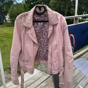 Rosa skinnjacka Jofama by Kenza - Snygg ljusrosa skinnjacka från Jofama by Kenza med coola detaljer som flätad axel, bälte i midjan och dragkedjor vid ärmslut. Jackan har klassisk bikerstil och blommigt foder på insidan. Perfekt för dig som vill sticka ut med en feminin touch.