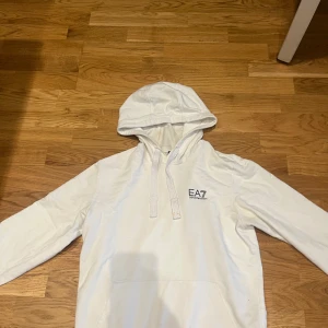 Vit hoodie från EA7 Emporio Armani - Snygg vit hoodie från EA7 Emporio Armani med logga på bröstet. Tröjan har huva med snörning, känguruficka och långa ärmar. Tillverkad i mjuk bomull som känns skön mot huden. Perfekt för en chill och stilren look. Xs/S