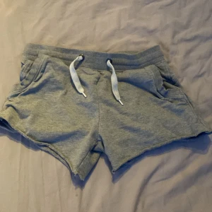 Grå mjuka shorts med snörning - Superbekväma grå shorts i mjukt bomullsmaterial med bred resår i midjan och vit snörning framtill. Perfekta för chill dagar eller träning. De har två fickor på sidorna och en avslappnad passform. Enkla att matcha med en hoodie eller t-shirt.