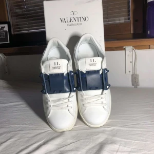 Valentino Rockstud Untitled sneakers vit/blå - Snygga Valentino Garavani Rockstud Untitled sneakers i vitt skinn med blått band och ikoniska nitar. Klassisk låg modell med rund tå, vita snören och platt sula. Perfekt för dig som vill sticka ut med exklusiva detaljer och modern design.