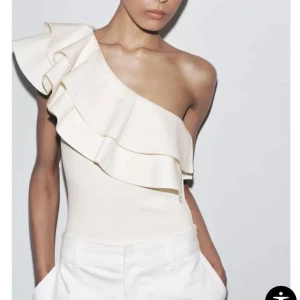 Vit one shoulder volangbody Zara - Superfin vit body från Zara med one shoulder och stora volanger över axeln. Sitter tight och har en snygg asymmetrisk form som verkligen sticker ut. Perfekt att styla med jeans eller kjol för en trendig look.