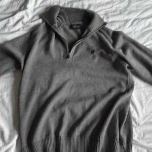 Grå stickad tröja Peak Performance - Snygg grå stickad tröja från Peak Performance med halv dragkedja och hög krage. Tröjan har diskret broderad logga på bröstet och ribbade muddar. Perfekt för lager-på-lager under kyliga dagar.