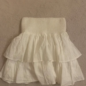 Vit volangkjol från KappAhl - Supersöt vit kortkjol från KappAhl med bred smock i midjan och dubbla volanger. Kjolen har broderade små hålmönster och är gjord i bomull, perfekt för en fräsch och somrig look.🌸