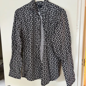 Svart blommig skjorta från Riley - Svart skjorta från Riley med vitt blommigt mönster och super slim fit. Skjortan har långa ärmar, klassisk krage och knäppning framtill. Perfekt för dig som gillar en smalare passform och vill sticka ut med ett snyggt mönster. Köptes på brothers för 900kr