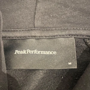 Svart hoodie från Peak Performance M - Svart hoodie från Peak Performance i storlek M. Klassisk modell med huva och snörning, stor magficka framtill och vit logga över bröstet. Perfekt för chill dagar eller när du vill ha en clean och enkel stil.