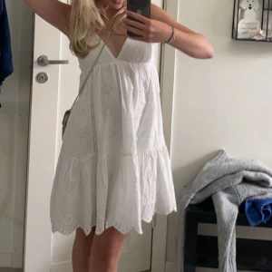 Vit somrig kortklänning med broderi - Superfin vit kortklänning med tunna axelband, v-ringning och broderade detaljer. Klänningen har volangkant nertill och snörning i ryggen för en söt och luftig look. Klänningen är från Zara och är i super bra skick🥰