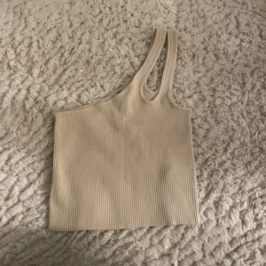 Beige ribbad oneshoulder topp - Trendig beige ribbad topp med oneshoulder-design och dubbla smala axelband på ena sidan. Perfekt för dig som gillar en clean och stilren look. Toppens material är stretchigt och sitter snyggt på kroppen. Den är en storlek M men funkar på dom som har S också.