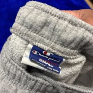 Grå mjukisbyxor från Champion - Grå mjukisbyxor från Champion med klassisk logga i midjan. Byxorna har resår och snörning i midjan för skön passform. Tillverkade i mjukt bomullsmaterial, perfekta för chill eller träning. Enkel och stilren design som funkar till allt.